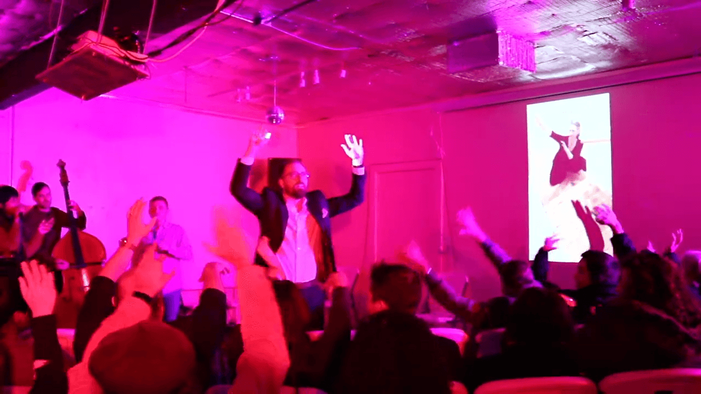 A performer with raised arms in front of the audience under magenta light, a projected dancer on the screen behind